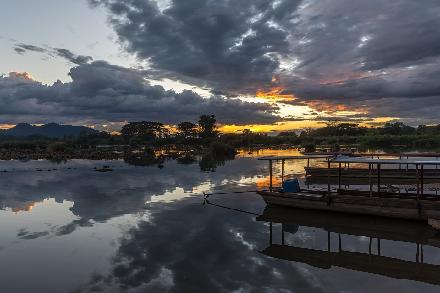 Sunset over the Mekong at Don Det, Laos: grey and orange clouds reflected in still water beside moored wooden boats.