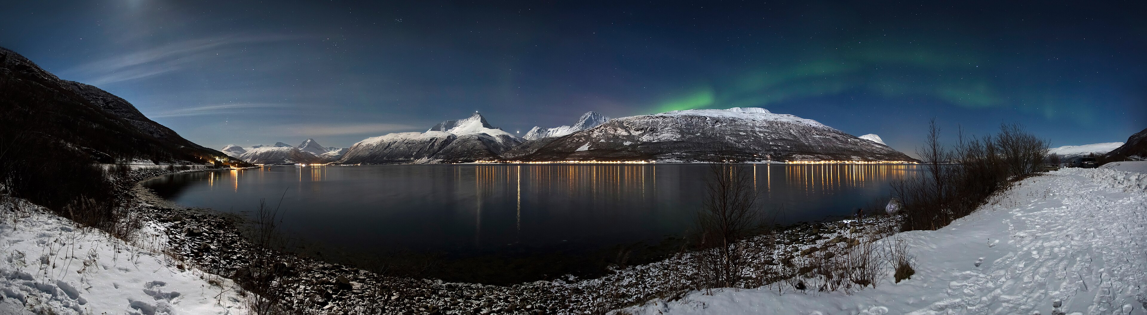 A wide winter panorama over Storfjorden in Norway: northern lights rippling above snowy mountains, village lights reflected in the still fjord.