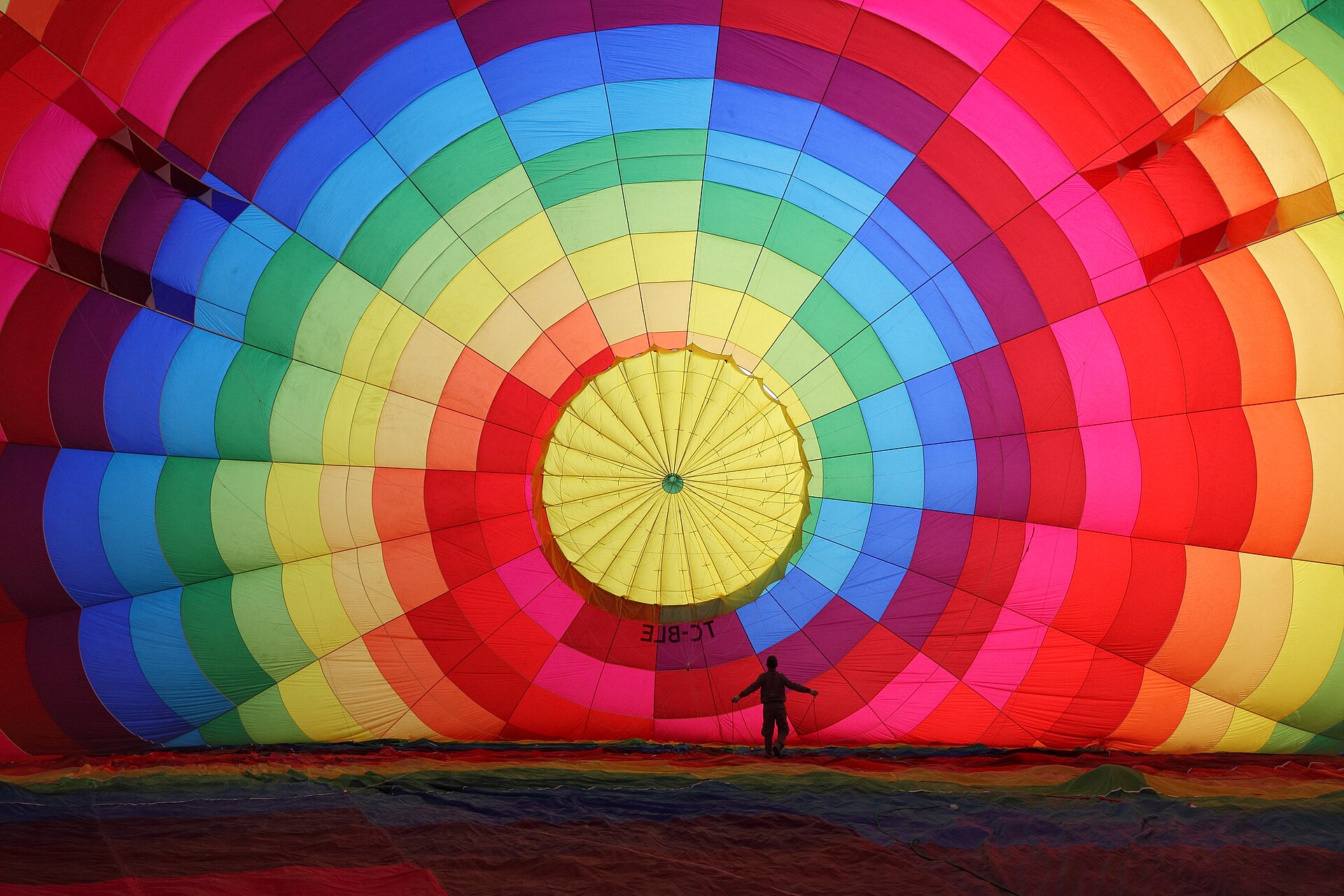 Inside a hot air balloon being inflated in Cappadocia: concentric rings of rainbow fabric rising to a glowing yellow crown, a person silhouetted at the centre holding the lines.