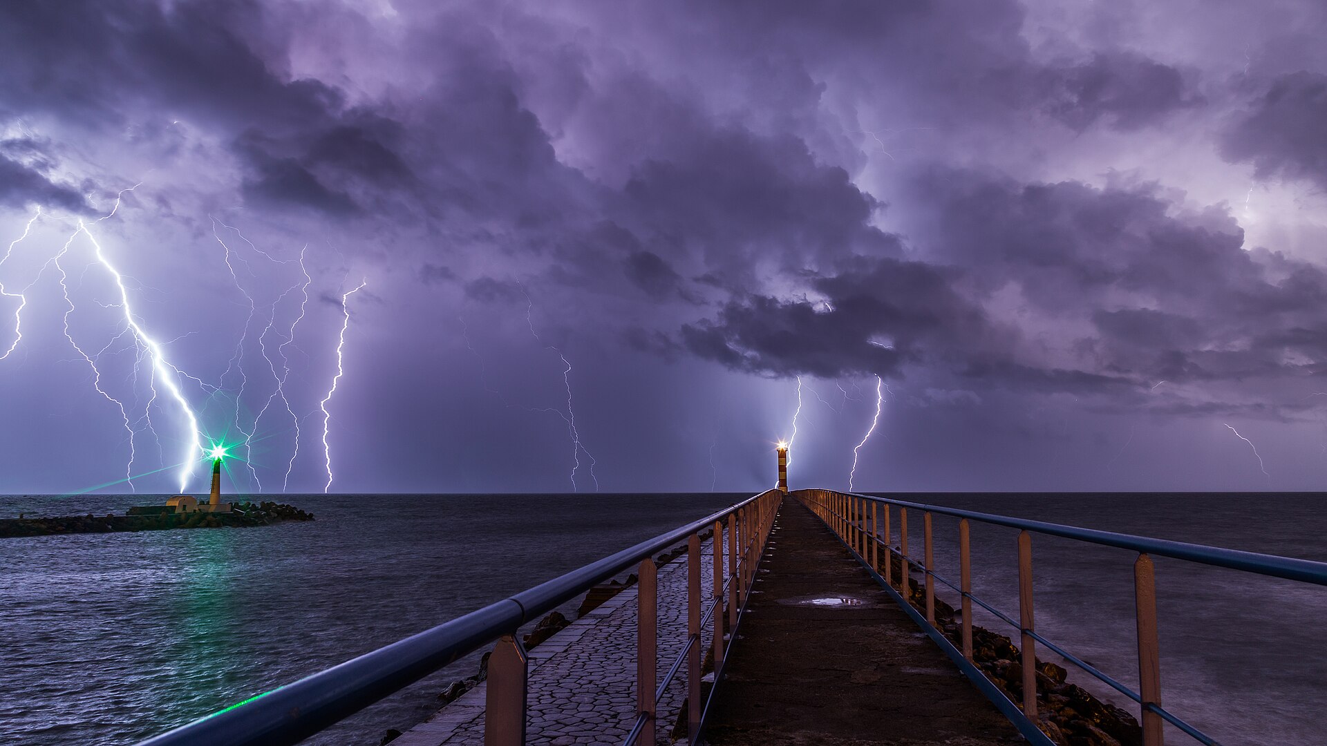 Lightning forking through a purple night sky over the harbour at Port-la-Nouvelle, two small lighthouses glowing green and gold at the end of a long pier.